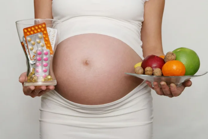 Kobieta w ciąży trzyma w jednej ręce owoce pełne witamin, a w drugiej tabletki z suplementami.